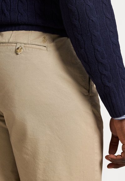 Μπεζ βαμβακερό παντελόνι με διακριτική υφή, που διαθέτει κλείσιμο με κουμπί και κεντημένο λογότυπο στην πίσω τσέπη. Συνδυάζεται με σκούρο μπλε πουλόβερ με κανονική πλέξη.