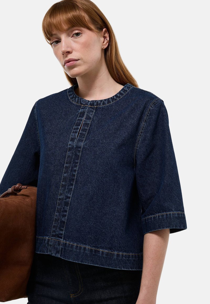 Chemise en denim bleu foncé avec un col rond, des manches trois-quarts et un détail de couture à l'avant. Assortie avec un sac marron.