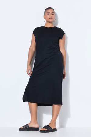 Mujer con vestido midi negro sin mangas y sandalias planas negras posando frente a un fondo gris claro.