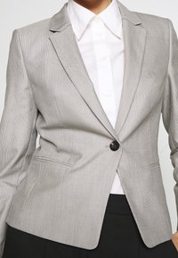 Blazer à carreaux gris avec revers crantés, fermeture à un bouton et deux poches avant. Porté avec une chemise blanche à col en dessous.