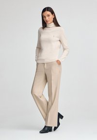 Maglione a collo alto beige realizzato in tessuto morbido, abbinato a pantaloni beige su misura con piega, e stivaletti neri con tacco.