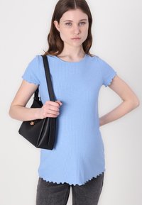 Tricou ribbed de culoare albastru deschis, cu mâneci scurte, cu tiv ondulat, și o geantă de mână de culoare neagră, cu un design minimalist, ținută în față.