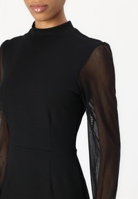 Svart klänning med hög hals och genomskinliga svarta prickiga ärmar. Tyget är slätt med strukturerade sömmar och en figursydd silhuett.