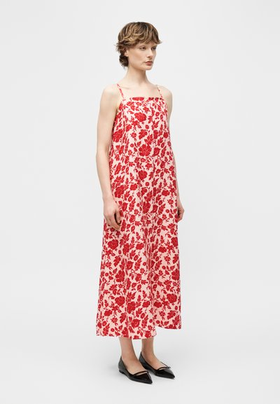 Γυναίκα που φοράει αμάνικο φόρεμα μήκους μέχρι τους αστραγάλους με κόκκινο λουλουδάτο σχέδιο σε ανοιχτό φόντο και μαύρες μυτερές μπαλαρίνες, στέκεται πλάγια.