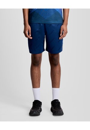 Man in marineblauwe sportshorts, witte enkelsokken en zwarte hardloopschoenen, staand tegen een effen lichtgrijze achtergrond.