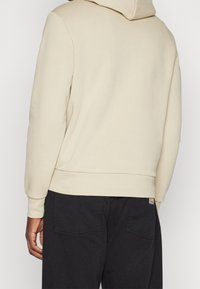 Felpa beige con cappuccio, dalla texture liscia, con polsini aderenti e tasca a marsupio, abbinata a pantaloni scuri con logo a patch.