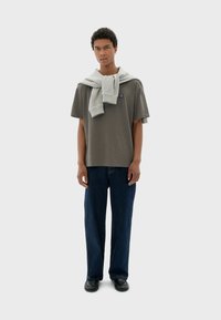 Chemise gris à manches courtes, jean en denim bleu ample, et chaussures noires, avec un pull gris clair drapé de manière décontractée sur les épaules.