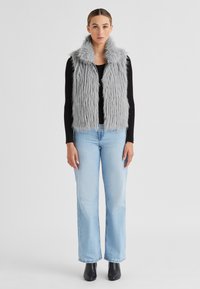 Graue Kunstfellweste mit flauschiger Textur und weitem Kragen, kombiniert mit einem schwarzen Langarmoberteil und hellblauen Schlagjeans. Schwarze knöchelhohe Stiefel.