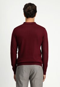 Burgundowy sweter z dzianiny z kołnierzykiem, dopasowany krój, długie rękawy. W połączeniu z szarymi spodniami z tylnymi kieszeniami.