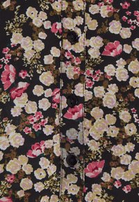 Tissu noir à motif floral dense avec des fleurs crème, roses et vertes, présentant une rangée verticale de trois boutons noirs cousus le long du centre.