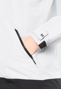 Lyst grå jakke med en lynlåslomme på ærmet, elastisk manchet og en subtil "SD" mærkebeskrivelse på manchetten.