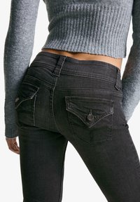 Black denim jeans with a mid-rise fit, featuring back pockets with pointed flaps and metal buttons. Worn with a light gray, ribbed knit sweater.