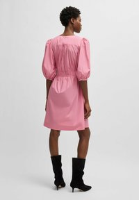 Pink puff-sleeve dress with cinched waist, knee-length. Smooth fabric, minimal detail on back. Model wears black knee-high boots.
