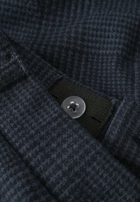 Marineblauer Stoff mit einem strukturierten Hahnentrittmuster, versehen mit einem transparenten Knopf und einem schwarzen Stoffakzent. Nahaufnahme zeigt die Nähdetails.