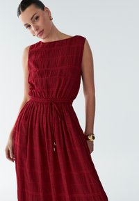 Rød, ærmeløs kjole med tekstureret stof med vandrette striber, elastisk talje og snøre-detalje.