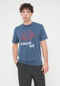 Μπλε βαμβακερό T-shirt με γραφικό καβούρι σε κόκκινο χρώμα και το κείμενο "PINCH ME" από κάτω. Λαιμόκοψη crew, κοντά μανίκια, άνετη εφαρμογή.