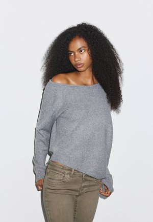 Femme aux cheveux bouclés portant un pull gris épaules dénudées et un pantalon vert olive, debout devant un fond blanc uni.