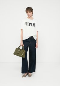 Jovem com t-shirt branca "REPLAY", calças largas escuras, saltos bejes, segurando uma mala verde-azeitona com detalhe em estrela.