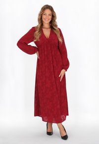 Vestido maxi rojo con mangas largas, escote en V profundo, detalle fruncido debajo del busto, patrón floral texturizado y silueta fluida. Combinado con tacones negros.