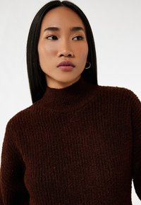 Brun højhalset sweater lavet af struktureret strikstof med ribbede detaljer, der har lange ærmer og en tætsiddende silhuet.