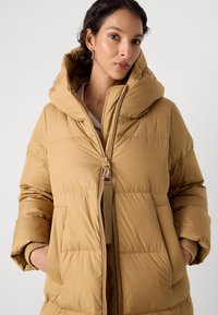 Béžová péřová bunda s vysokým golierom a oversized kapucňou. Obsahuje centrálny zips, horizontálne prešívanie a bočné vrecká. Hladká textúra.