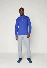 Camisola atlética de manga longa azul com colarinho de meia abertura, combinada com calças cinzas claras e ténis azuis. Tecido liso e elástico.