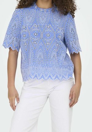 Vrouw draagt een lichtblauwe kortärmelig kanten blouse met geometrische patronen, gecombineerd met witte broek en gouden ringen aan haar vingers.