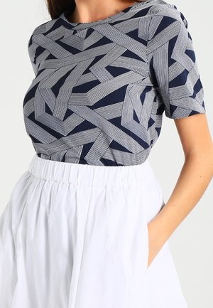 Femme portant un haut à manches courtes à motif géométrique bleu marine et blanc, rentré dans une jupe blanche taille haute avec des poches.