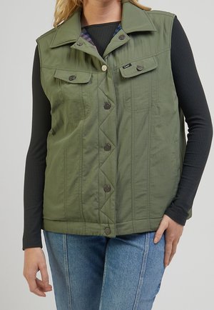 Kobieta ubrana w zieloną kamizelkę na guziki, na czarnym prążkowanym bluzce z długim rękawem i niebieskich dżinsach, stojąca z jedną ręką swobodnie opuszczoną, a drugą na biodrze.