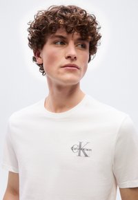 Νεαρός με καστανά σγουρά μαλλιά που φοράει λευκό μπλουζάκι Calvin Klein, κοιτάζοντας προς τα πλάγια μπροστά σε απλό φωτεινό φόντο.