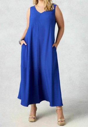 Vrouw draagt een mouwloze koningsblauwe maxi-jurk met zakken en beige sleehak sandalen op een lichtgrijze achtergrond.