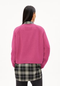 Pull rose en tricot avec manches raglan, présentant un panneau arrière texturé, porté par-dessus une chemise à carreaux noire et verte.