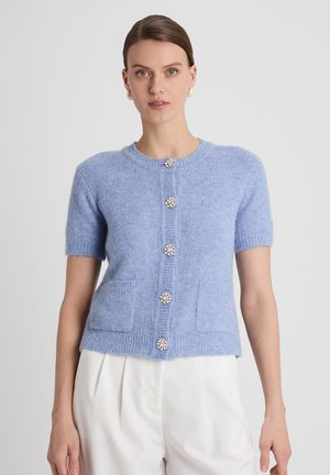 Woman wearing a light blue short-sleeve cardigan with decorative buttons, paired with white high-waisted trousers, standing neutral.