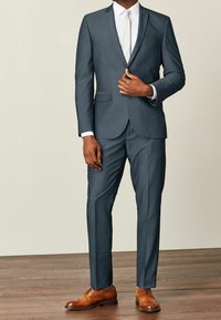 Mężczyzna w ciemnoszarym garniturze z białą koszulą, białym krawatem i brązowymi eleganckimi butami, stoi na drewnianej podłodze na jednolitym tle.