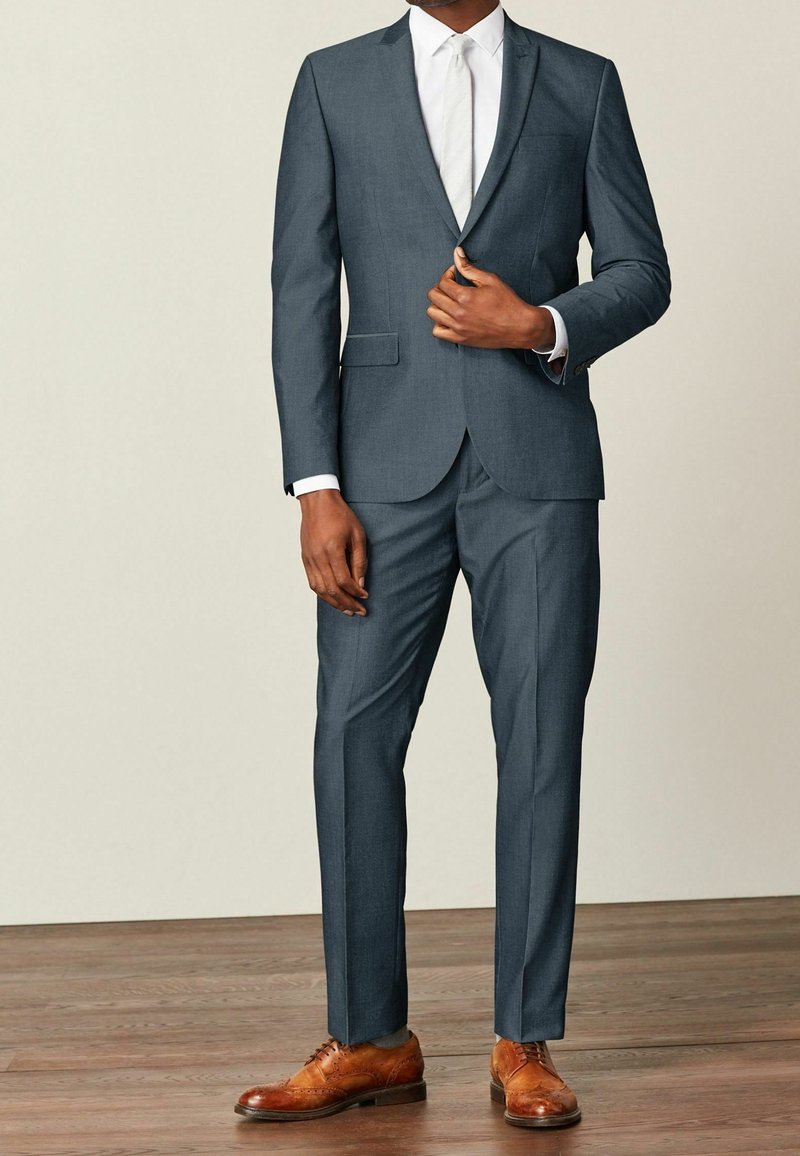 Mężczyzna w ciemnoszarym garniturze z białą koszulą, białym krawatem i brązowymi eleganckimi butami, stoi na drewnianej podłodze na jednolitym tle.