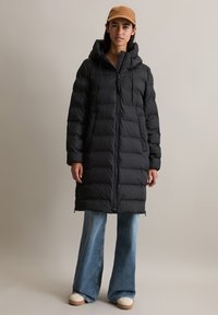 Svart dunjakke med hette, med glidelås foran og snøring. Kombinert med utslåtte denimbukser og hvite joggesko.