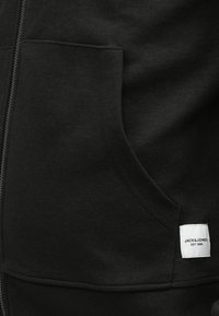 Sweat à capuche noir zippé avec poche kangourou à l'avant et étiquette rectangulaire blanche près de l'ourlet portant l'inscription « JACK & JONES EST. 1990 ».