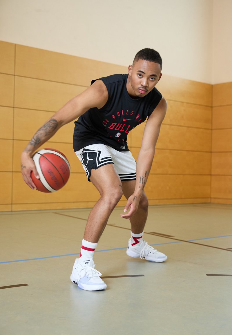 Joueur de basketball en débardeur noir, portant un short à motifs blancs et noirs, des baskets blanches, dribblant un ballon de basket rouge sur le sol du gymnase.