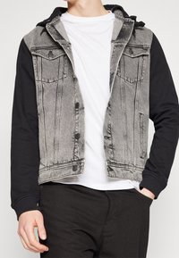 Person som bär en svart och grå denimjacka med svarta ärmar, vit t-shirt och svarta byxor med ena handen i fickan.