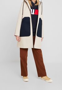 Béžový voděodolný kabát s námořnickými kapsami, kapucí a červenými detaily. Nosený přes tmavý svetr a hnědé kalhoty, doplňuje ležérní vzhled.