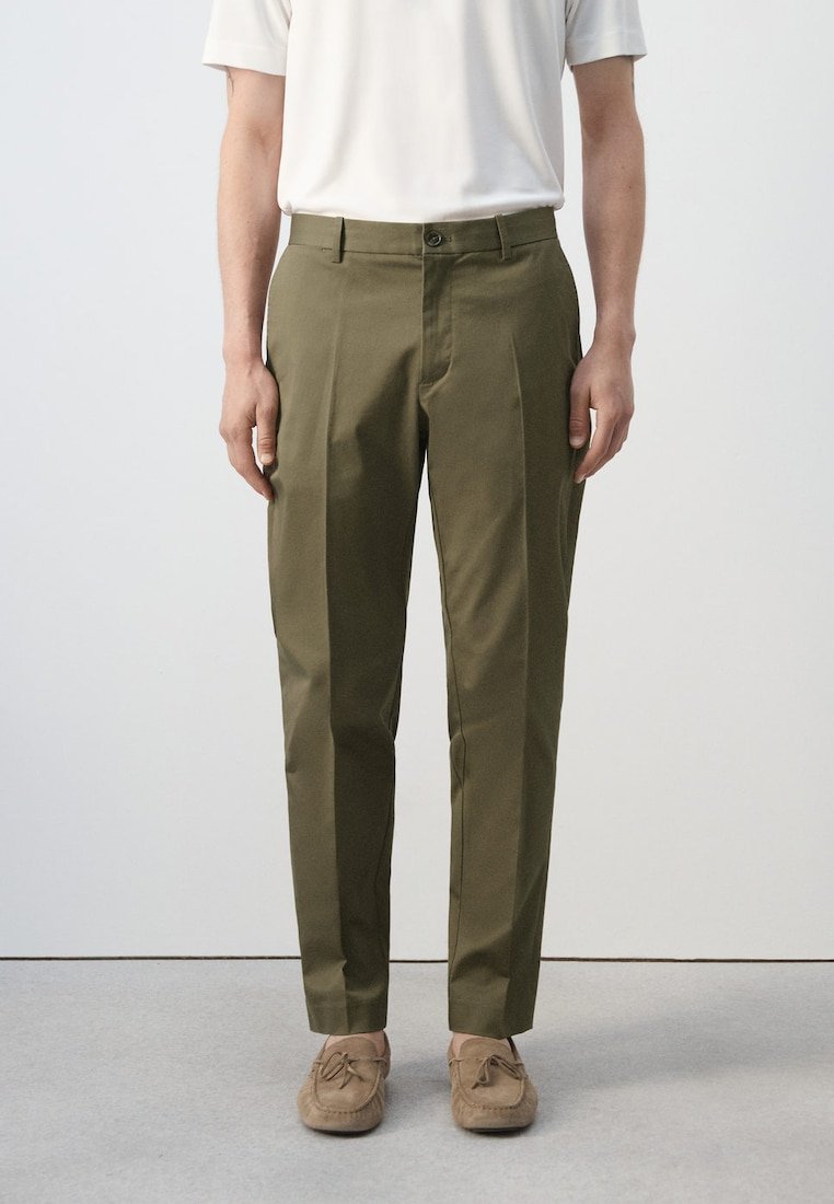 Homme portant un pantalon vert olive sur mesure, des mocassins beiges et une chemise blanche à manches courtes, debout devant un fond uni.