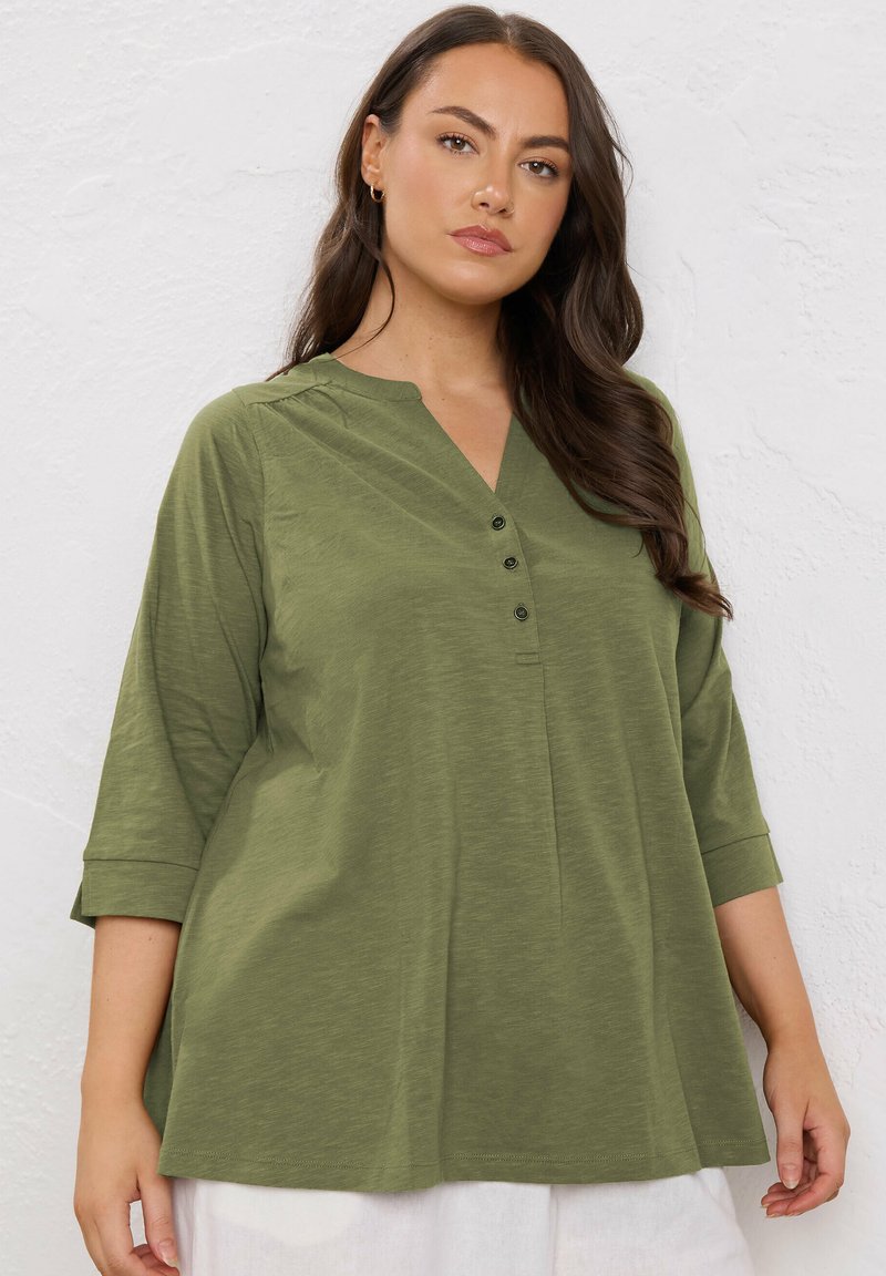 Donna con lunghi capelli castani che indossa una camicetta verde oliva con bottoni e maniche a tre quarti, davanti a un muro bianco e testurizzato.