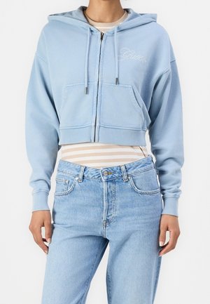 Osoba nosząca jasnoniebieską krótką bluzę z kapturem na zamek i kieszeniami z przodu na beżowo-białym paskowanym topie, w połączeniu z jasnoniebieskimi jeansami z wysokim stanem.