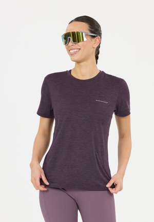 Frau lächelt, trägt ein dunkelviolettes Sportshirt, passende Leggings und große reflektierende Sportsonnenbrille, steht vor weißem Hintergrund.