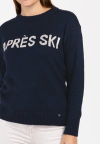Marineblauer Strickpullover mit dem Text "APRES SKI" in weißen Buchstaben. Gerippter Ausschnitt und Bündchen, mit einem kleinen metallischen Logopatch.