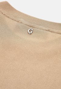 Vaaleanbeige puuvillasekoitekangas yläosa, jossa on uritettu tekstuuri ja hopeinen 'G'-logo kaula-aukossa. Yksinkertainen muotoilu pyöreällä kauluksella.