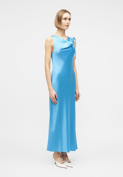 Γυναίκα με κοντά ξανθά μαλλιά, ντυμένη με αμάνικο, ανοιχτό μπλε σατινέ φόρεμα και άσπρες μυτερές γόβες με λουριά στον αστράγαλο, που στέκεται μπροστά σε απλό φόντο.