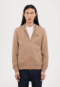 Bež zip-up dukserica s kapuljačom na vezanje, kenguru džepovima, rebrastim rukavima i EA7 logom na lijevoj strani prsnog koša. Nosiva preko bijele majice.
