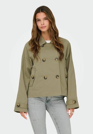 Hosszú hullámos hajú nő, aki olíva zöld kétsoros gombos zakót és világosszürke farmert visel, egyszerű háttér előtt áll.