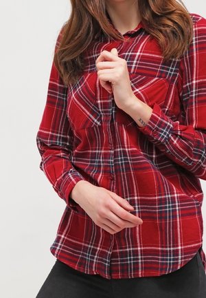 Red plaid flannel shirt with button closure, long sleeves, and two chest pockets. Features white and navy check patterns on a textured fabric.